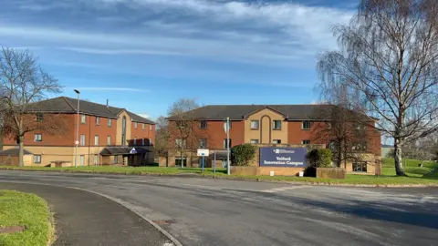 The exterior of a building at the University of Wolverhampton's Telford campus. The picture is taken from the opposite side of a road near the building, showing the red-brick building with University of Wolverhampton signage, and trees and bushes around the building.