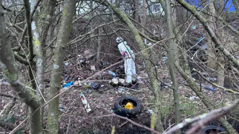 Kate Bradbrook/BBC A person in a protective suit works on a clearup of waste on a woodland verge close to a road. 