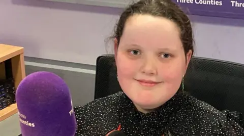 Justin Dealey/BBC Betsy Griffin is in the BBC 3CR radio studio sitting in a chair in front of the purple microphone. She is smiling at the camera and is wearing a sparkly black top.