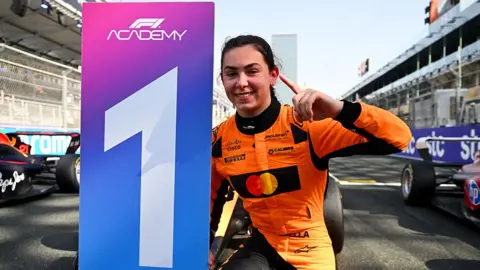 Getty Images Ella Lloyd, wearing an orange race suit with black sponsor logos, holding up a "one" sign with her fingers while crouching next to a "one" winners board. She is on the race track in front of a Formula 1 car.