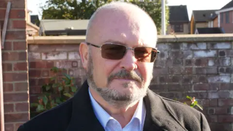 John Fairhall/BBC Graham Plant is wearing dark tinted sunglasses and is looking at the camera. He is standing in front of a brick wall and is wearing a striped blue shirt and black coat. 