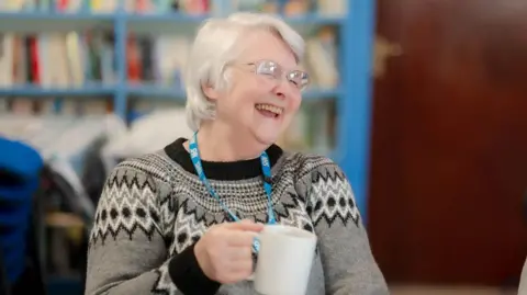 A lady laughs as she holds a tea mug. She has grey hair and is wearing glasses and a grey patterned jumper.