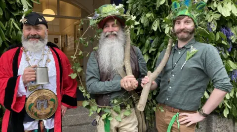 Mock Mayor and men of the woods at Flora Day. The three men are standing in a row and are dressed up. The man on the left is wearing a black hat, red robe and is holding a silver tankard. The man in the middle has a long beard and is wearing an ivy covered hat. The man on the left also has green foliage on his hat.
