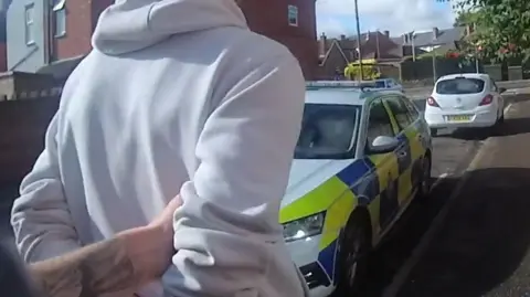 Image of Lakaiko after being arrested with a police car in the background. 
He is wearing a white hooded sweatshirt and his arms are restrained with handcuffs visible on his wrists.  