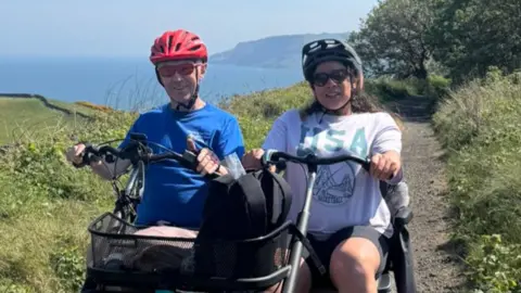 A man and woman are on a cycle track wearing cycle helmets. They are riding an adapted bicycle sitting next to each other.