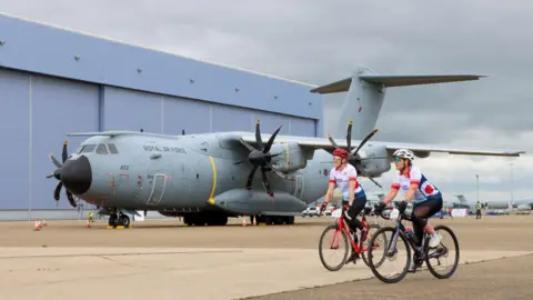 Two cyclists on bikes near a RAF plane. Other planes can be seen in the distance. It is a cloudy day.