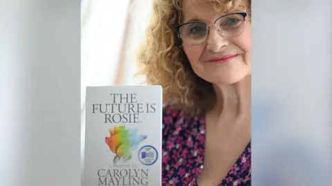Carolyn Mayling stood holding her book and looking to the camera