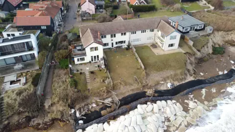 John Fairhall/BBC An aerial image of the Thorpeness coastline. It shows a large home on the edge of the cliff and piles of large white sandbags acting as sea defences. 
