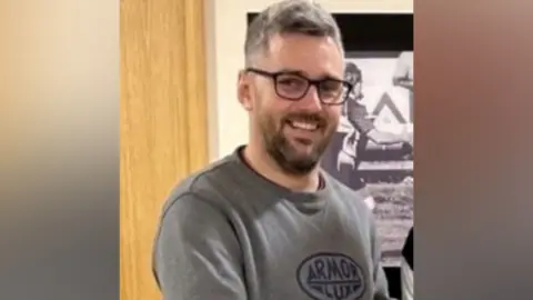 BBC Craig Hurcombe is wearing glasses, has short greying hair and has facial hair. He is smiling at the camera.