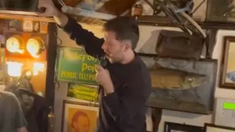 A man in a black jumper, stood in a bar, singing down a microphone