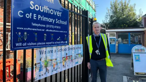 Headteacher Noel Fadden standing outside the front gates of Southover Primary School. He has a high visibility yellow jacket on.