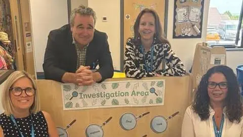 NHS Cornwall and Isles of Scilly ICB Staff members at Bugle school in Cornwall, accompanied by Dr Chris Reid, Chief Medical Officer, NHS Cornwall and Isles of Scilly ICB, pictured smiling at the camera in a school classroom, alongside a poster saying "Investigation Area" and pictures of magnifying glasses.