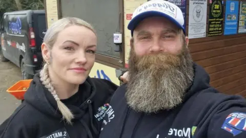 Higgsy's Amber wearing a black hoodie and Adam wearing a black hoodie, smiling for a selfie in front of their burger shed.