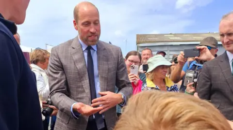 Sharon Hart Prince William, dressed in a grey patterned suit with a light blue shirt and navy blue tie with his hand out accepting a crisp. There are people in the background taking photos on their phones.