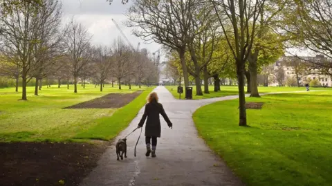 The back of a woman walking a dog in a park. She is on a footpath and there is grass either side of her. (generic image)