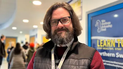 Matt Tyrer has brown hair which is straight and just below his ears in length. He wears glasses and has a beard. He is wearing a red jumper under a black gilet and standing inside with a group of people behind him.
