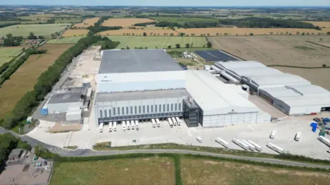 An aerial view shows a large industrial warehouse complex with multiple loading bays and lorries, surrounded by expansive farmland and fields.