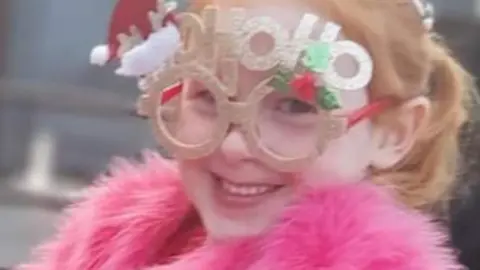 Family photograph Lila with red hair wearing Christmas glasses which say 'HoHoHo' and a pink fur jacket smiling.