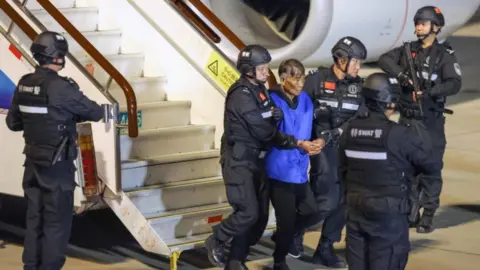 An elderly man in a blue vest being escorted off a plane by police with helmets.