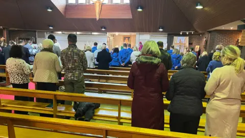 Richard Knights/BBC People dressed in coats in a church. They are standing with heads slightly bowed while a bishop in pink robes can be seen at the front of the church.