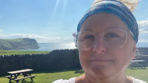 Romy Wood Romy Wood outside in picnic area on grass with tables behind and the sea in the background on sunny summer's day. She wears a white t-shirt and glasses and blue patterned bandana in her hair
