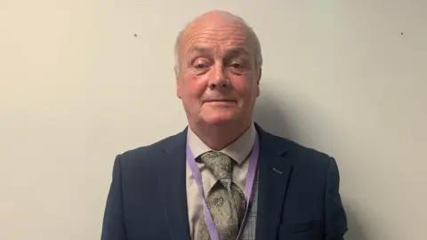 A bald man standing against a white wall. He is wearing a green tie and white shirt. He has a purple lanyard on and is looking at the camera.