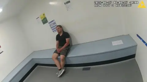 Kent Police A man sits in a police cell. The room is bare and white. 