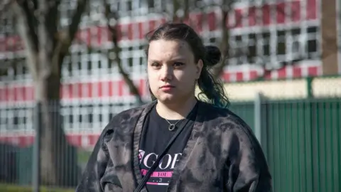 A young woman, with long dark hair tied up in a bun looks at the camera from outside a large school building. She wears a dark tie-dye cardigan and black t-shirt.