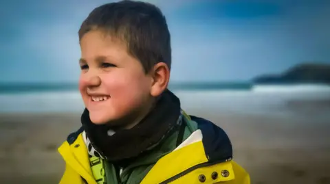 Family photo Ivor looks at an angle smiling. He has short light brown hair and wearing a yellow fisherman's jacket. He is standing on beach with surf and a point in the distance