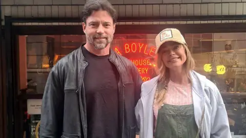 Aaron Phypers with brown hair and black leather jacket and black t-shirt with actress Denise Richards wearing a beige baseball cap patterned t-shirt, dungarees and grey hoodie standing outside Ma Boyle's eatery in Liverpool.