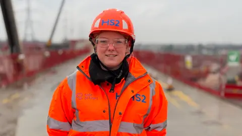 A woman in an orange high-visibility jacket, wearing an orange hard hat and protective goggles, standing in from the the viaduct building site