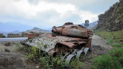 A rusted, wrecked tank lies on the side of a rural road. Mountains are visible in the background.