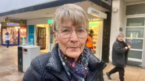 Helen Mulroy/BBC Image shows 78 year old Sue Lancaster, with grey-blonde hair and wearing a black puffer jacket and patterned scarf looking at the camera. She stands in front of shops in Bedford town centre. 