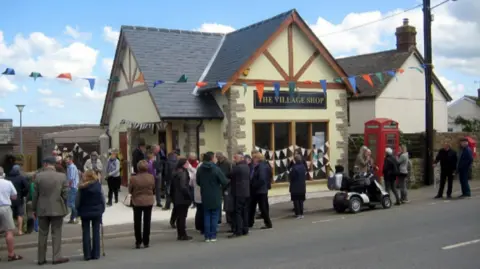 A magnolia building with a wooden-gabled roof, bunting in the windows, with a red post box next to it. A black and gold sign says The Village Shop and the shop is surrounded by villagers including one on a mobility scooter.