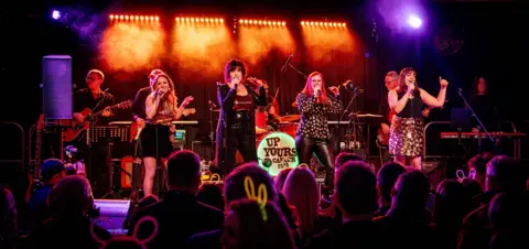 Jane Evans Four women in a line on stage. They are wearing gold and black outfits. Behind them are musicians and in front is the audience.