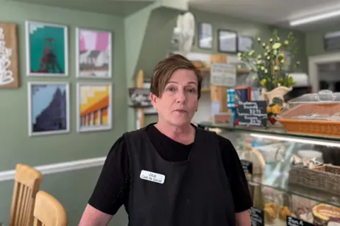 Julie, a woman in her 60s with short brown hair swept over her right ear, wearing a black t-shirt with black apron over and a name badge which reads Julie, Café  De Galles. She is standing in front of a café counter which has baked goods and baskets inside and a bunch of yellow carnations on top. There is an olive green wall behind to the left and artwork, out of focus. 