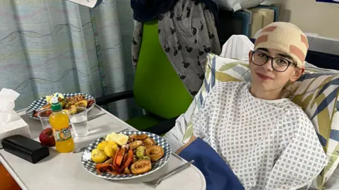 Family A boy with glasses in a surgical robe, with his head bandaged and a hospital tray in front of him with a large dinner on it