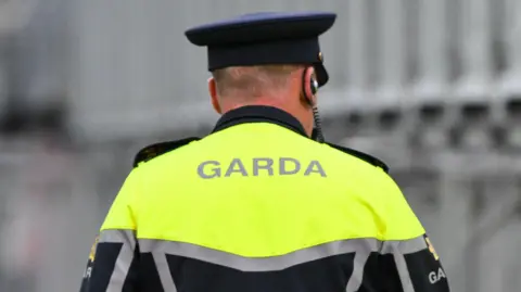 A garda (Irish police) office on patrol. The man, viewed only from behind, is wearing a navy peaked cap and a dark uniform with yellow high-viz shoulders and silver reflective stripes. The background is blurred. 