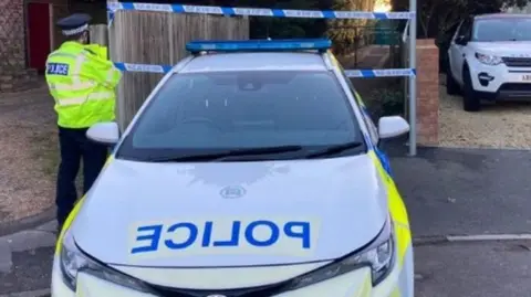 A marked police car is pictured parked in front of an alleyway which is cordorned off by police tape. A police officer is pictured beside the car looking towards the alley.