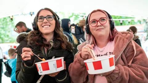 Headland Two women in glasses, one with brown hair, one blonde, smile as they hold up red and white KFC buckets while dipping their wooden forks inside.