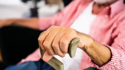 A close-up image of an elderly person's hand gripping the top handle of a walking stick. They appear to be sitting down while wearing a red and white chequered shirt over a white T-shirt.