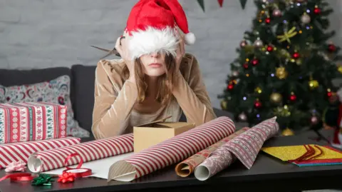 Woman with a Santa hat pulled down over her eyes sitting at a table with rolls of wrapping paper in front of her.