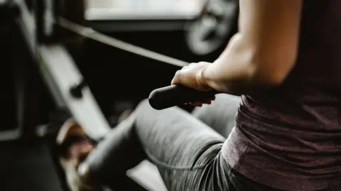 Getty Images A woman in gym clothing is seen on an indoor rowing machine.
She is wearing a light purple top and grey sport leggings.