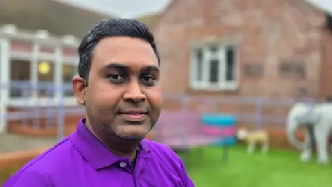 Paul Moseley/BBC Alamin Mia wearing a purple polo shirt. He is outside. The background is out of focus, but there appears to be a model of an elephant next to a one-storey brick building.