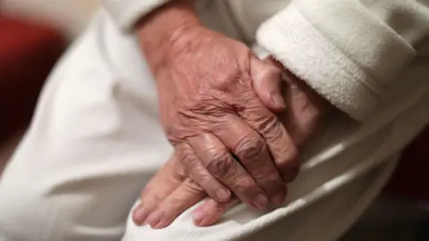 PA Media A close up of an elderly person's hands rest on white trousers. 