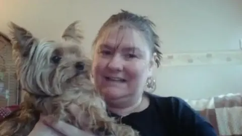 Ann Green is cuddling a pet dog and sitting on a sofa. She is wearing a black top and has her hair tied back in a ponytail and is wearing earrings.