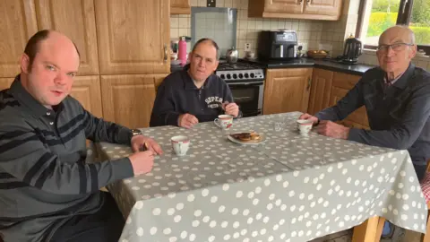 Alphie Lonergan sits at his kitchen table with his sons Ronan and Donal. Donal, in the middle, is wearing a navy quarter-zip top and has receding dark hair. Ronan is wearing a striped top and is bald on top with dark hair at the sides. The kitchen table has a grey/green tablecloth with white polka dots on it and there is a plate of biscuits in the middle. A mug is in front of each of the three men.