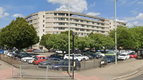 Google A large multi‑storey concrete building with long horizontal balconies stands behind a row of tall green trees. 