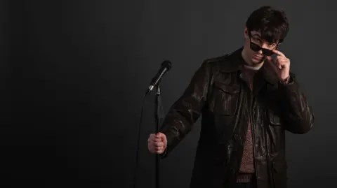 Burt Williamson Burt Williamson wearing a black leather jacket and holding a mic stand in one hand, while pulling his sunglasses down from his eyes with the other hand.