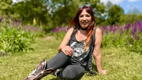 Martin Giles/BBC Justine Hatfield sits in a grass clearing surrounded by purple wildflowers. She wears black jeans, burgundy cowboy boots and a t-shirt which says 'The Flying Pig Stage, Strawberry Fair 2022.'